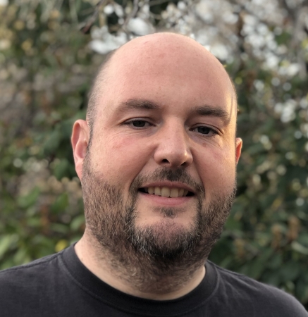 Smiling man with a beard and no hair in a tshirt with trees behind