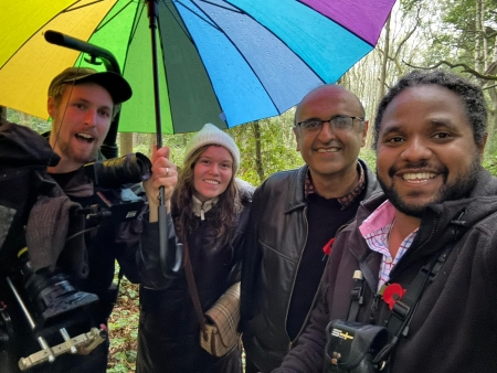 3 mena and a woman with a wolly hat on stand under a multi coloured umbrella