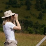 Woman with a hat on looking through binoculars, green hills behind
